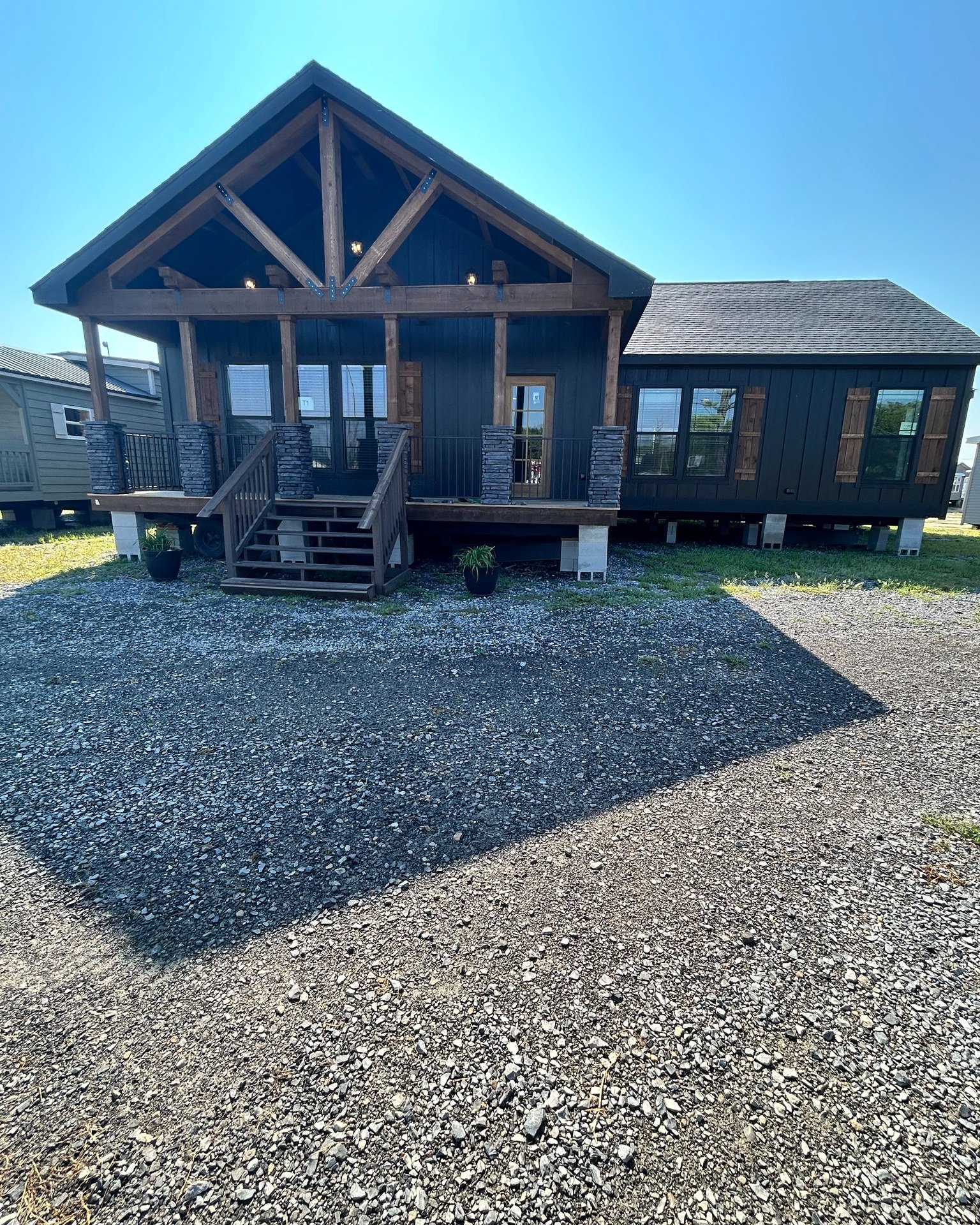 A modern cabin with a wooden porch and exposed beams under a clear blue sky. Gravel pathway leads to steps with potted plants. Calm and rustic.