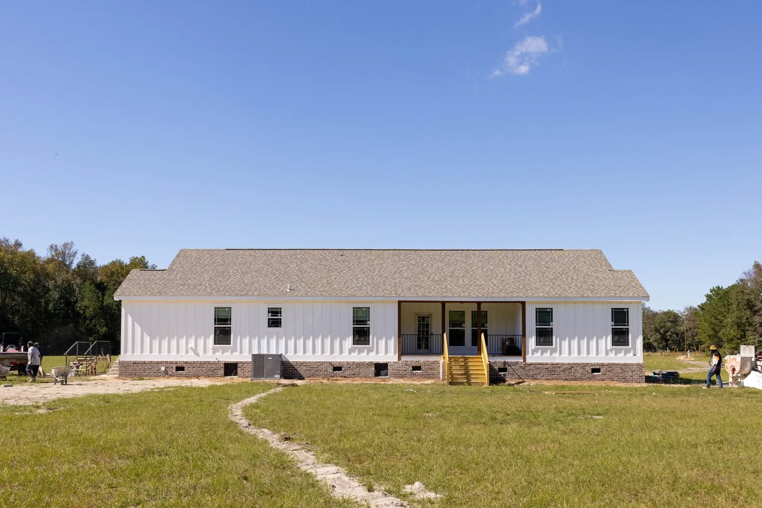 A single-story white house with a gray roof and yellow steps, set on a grassy lawn under a clear blue sky. A few people are visible near the home.