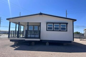 A modern modular home with a white exterior and dark trim sits on gravel. It features a shaded porch with railing and three front windows, under a clear blue sky.