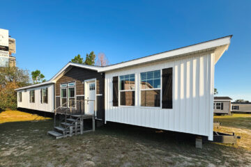 A white and brown modular home with a gabled roof sits on a grassy lot under a clear blue sky. Large windows and a small porch are visible.