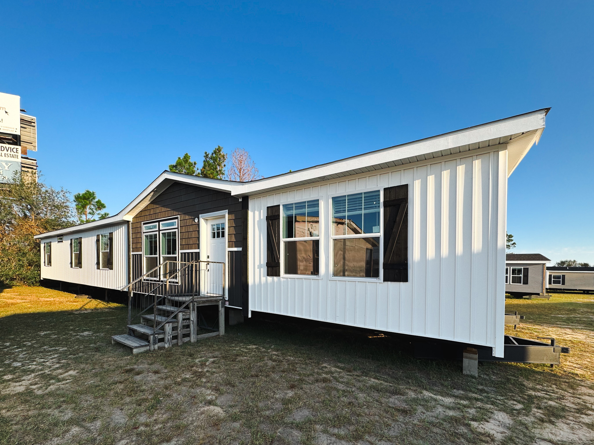 A white and brown modular home with a gabled roof sits on a grassy lot under a clear blue sky. Large windows and a small porch are visible.