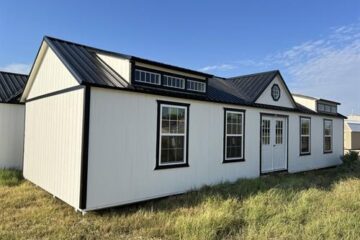 Empty room with freshly installed gray carpet partially covered by plastic, beige walls, and two windows providing natural light. A doorway leads to a white-tiled bathroom.