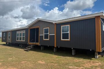 A modern modular home with dark gray siding and wood trim sits elevated on blocks with a cloudy sky in the background, conveying a sense of new beginnings.