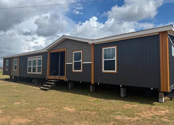 A modern modular home with dark gray siding and wood trim sits elevated on blocks with a cloudy sky in the background, conveying a sense of new beginnings.