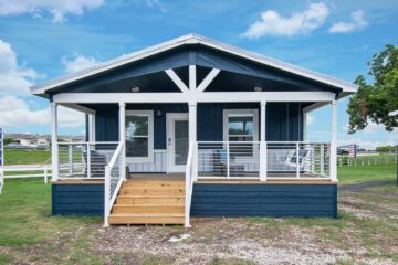 A small blue cabin with a white trim and covered porch sits on a grassy lawn under a bright blue sky. Wooden steps lead to the entrance, creating a cozy, inviting feel.