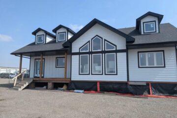 A modern white house with black trim features large triangular windows, dormer windows on the roof, and a small porch with stairs, set against a clear blue sky.