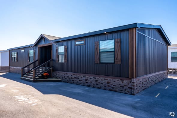 Modern manufactured home with dark siding and brick foundation. Wooden shutters frame the windows. A small porch with steps features potted plants.