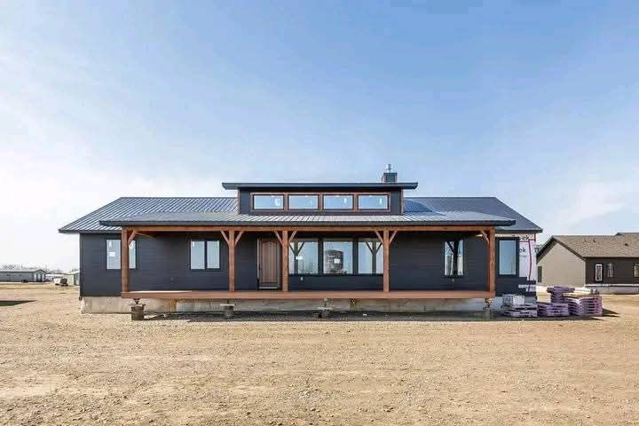 Single-story house with dark siding, a metal roof, and a long covered porch with wooden beams. The home sits on a large dirt lot under a clear blue sky.