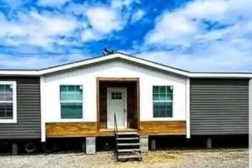 A mobile home with a gray exterior and white trim, set on a gravel lot. A few steps lead to the central door, under a cloudy blue sky.