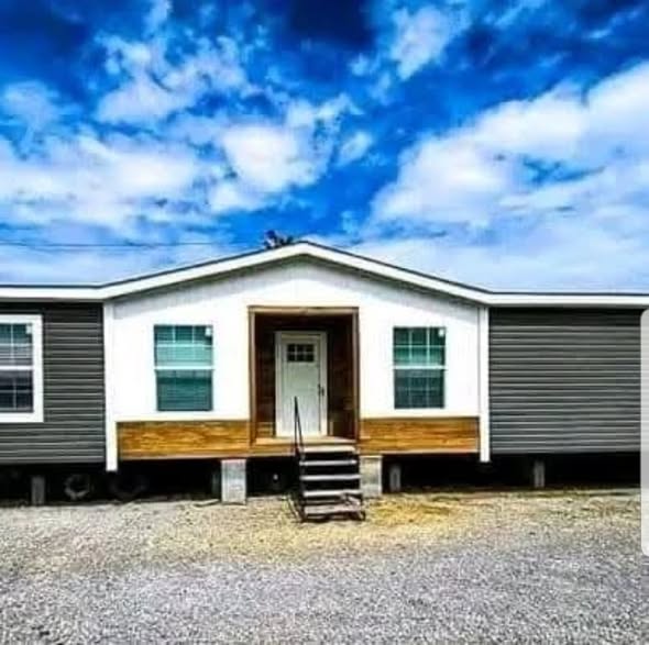 A mobile home with a gray exterior and white trim, set on a gravel lot. A few steps lead to the central door, under a cloudy blue sky.