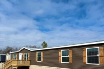 A long, brown manufactured home with wooden shutters and a small porch sits under a partly cloudy sky. The setting feels calm and open.
