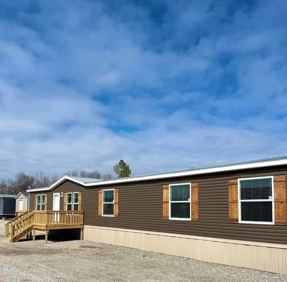 A long, brown manufactured home with wooden shutters and a small porch sits under a partly cloudy sky. The setting feels calm and open.