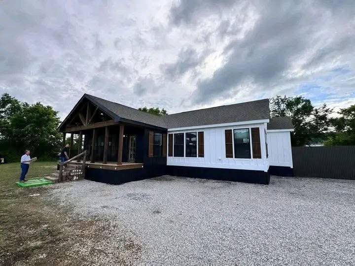 A modern, single-story house with white siding and a dark porch under a cloudy sky. Two people stand near the porch, and trees are in the background.