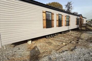 A beige mobile home is set on a gravel lot under a clear sky. Wooden shutters and large windows line its side. Trees are visible in the background.