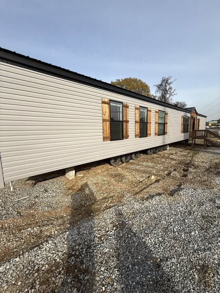 A beige mobile home is set on a gravel lot under a clear sky. Wooden shutters and large windows line its side. Trees are visible in the background.