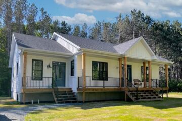 A modern, single-story white house with a dark roof, fronted by a porch with wooden pillars and black railings. It's set against a backdrop of tall trees and a blue sky.