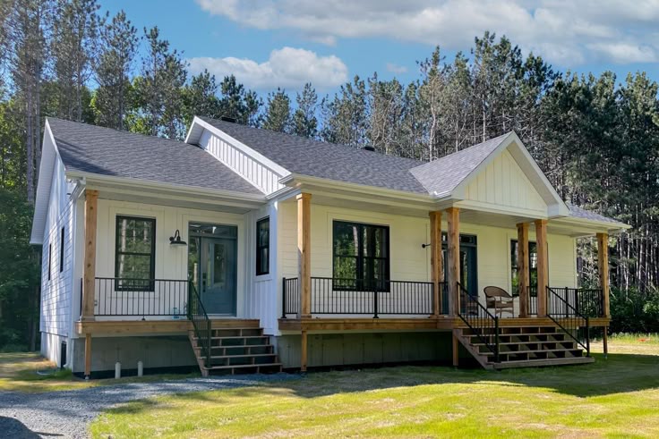 A modern, single-story white house with a dark roof, fronted by a porch with wooden pillars and black railings. It's set against a backdrop of tall trees and a blue sky.