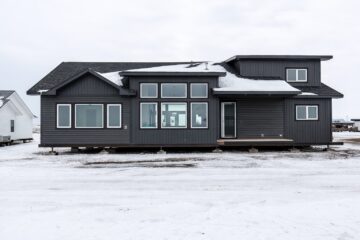 Modern dark gray house on a snowy landscape with large windows and white-trimmed roof. The scene is serene and wintry under an overcast sky.