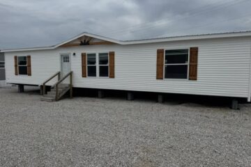 A white mobile home with wooden shutters and a small porch sits on a gravel lot under a cloudy sky, conveying a simple, rustic atmosphere.