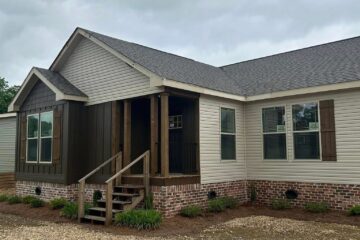 Single-story house with gray siding, dark brown accent, and brick foundation. Gravel driveway and small porch under cloudy skies, creating a calm mood.
