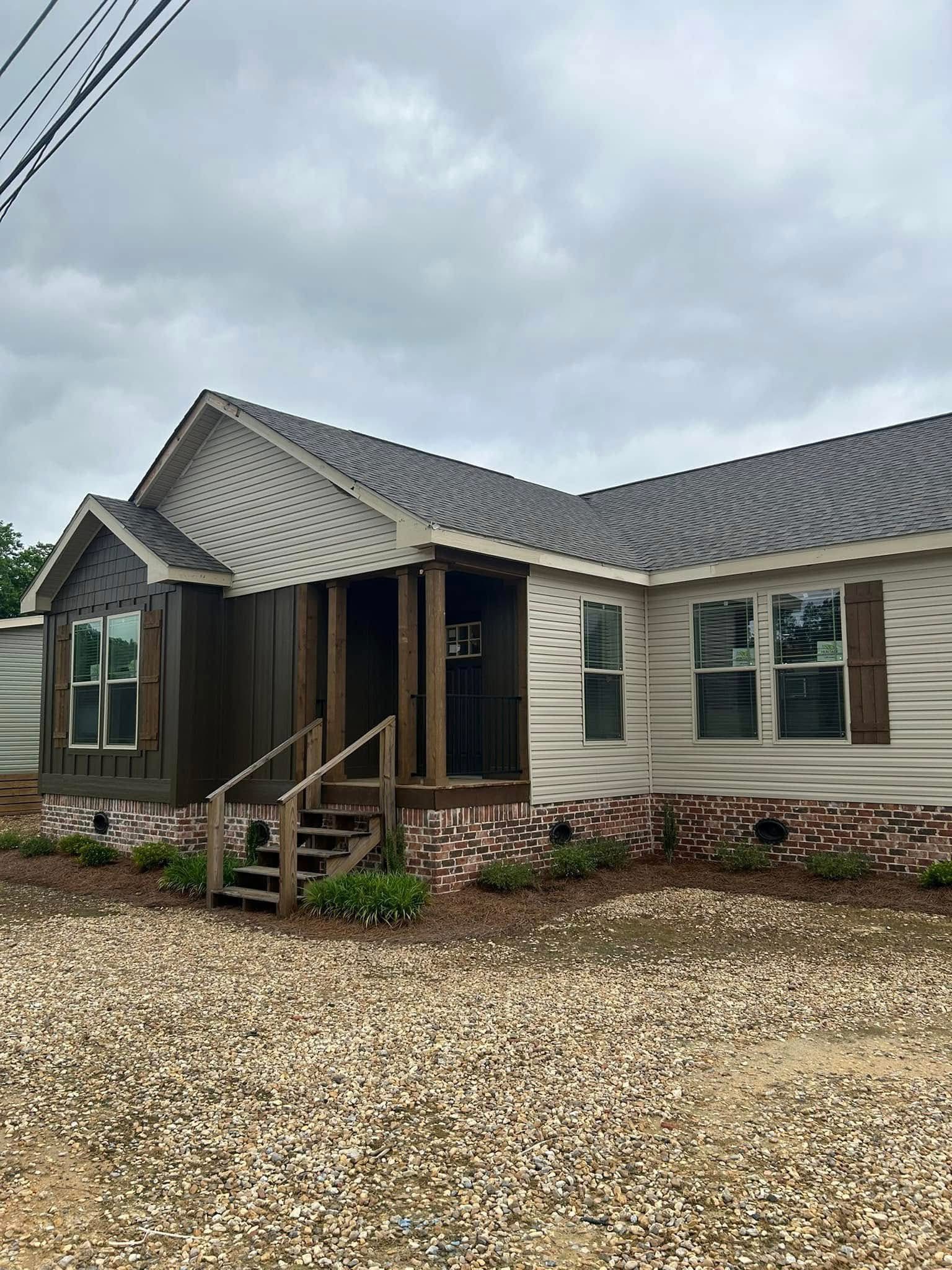 Single-story house with gray siding, dark brown accent, and brick foundation. Gravel driveway and small porch under cloudy skies, creating a calm mood.