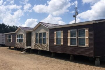 Manufactured home with four bedrooms and two bathrooms. Features brown siding and stone accents, set against a clear sky with fluffy clouds.