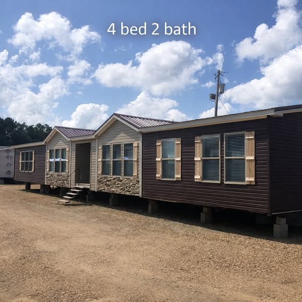 Manufactured home with four bedrooms and two bathrooms. Features brown siding and stone accents, set against a clear sky with fluffy clouds.