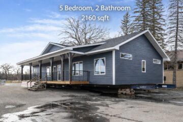 A blue modular home is placed on a flatbed trailer, featuring a porch, large windows, and a gabled roof. Text above reads: “5 Bedroom, 2 Bathroom, 2160 sqft.”