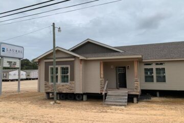 A beige modular home with stone accents sits on a dirt lot under a cloudy sky. A sign reads "Laurel Home Center, Summer Sale, Open House, July 10-12."
