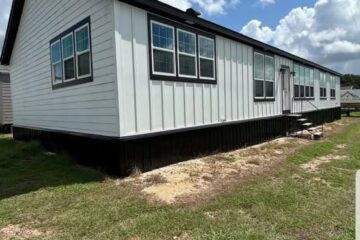 White manufactured home with black trim, multiple large windows, and steps leading to the entrance. Set on grassy land under a partly cloudy sky.