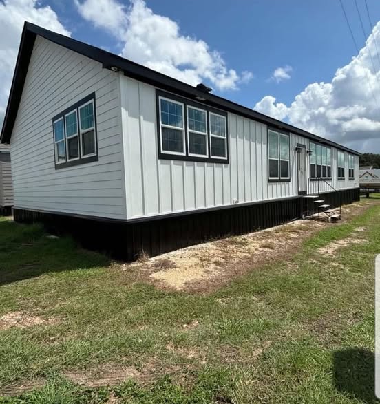 White manufactured home with black trim, multiple large windows, and steps leading to the entrance. Set on grassy land under a partly cloudy sky.