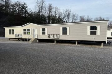 A beige mobile home with "5 Bed 3 Bath" and "$63,900" displayed on the side. It's set in a gravel lot, surrounded by leafless trees under a cloudy sky.