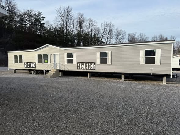A beige mobile home with "5 Bed 3 Bath" and "$63,900" displayed on the side. It's set in a gravel lot, surrounded by leafless trees under a cloudy sky.