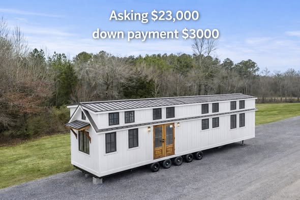 A long white tiny house on wheels sits on a gravel path, surrounded by grass and trees. Text above reads "Asking $23,000, down payment $3000."