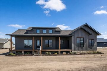 Modern farmhouse with gray siding, large windows, and gabled roof under a blue sky. Wooden porch supports add warmth, surrounded by a sparse landscape.