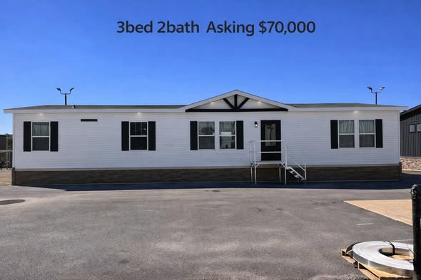 A manufactured home with a white exterior, black shutters, and a small porch. Text above reads "3bed 2bath Asking $70,000" against a clear blue sky.