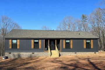Single-story dark gray house with wooden shutters, set against a clear blue sky and sparse wooded area. Text reads "3 bd, 2 ba, 1,568 sqft."