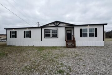 Single-story manufactured home with white siding and black trim sits on a gravel lot. The overcast sky creates a calm, muted atmosphere.