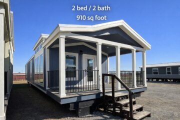 Small blue manufactured home with white trim and a covered porch. Sign reads "2 bed / 2 bath, 910 sq foot." Clear sky and additional homes in background.
