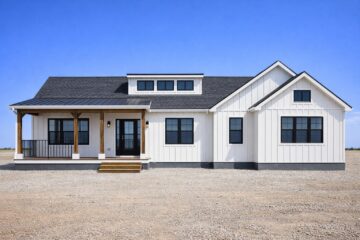 Modern white farmhouse with a black roof and wooden porch set against a clear blue sky. Symmetrical design conveys a clean, serene ambiance.