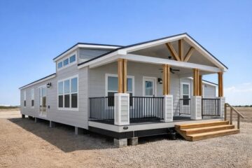 A modern, prefabricated home with gray siding and large windows, featuring a spacious front porch with wooden pillars and steps, set on a barren landscape under a clear blue sky.