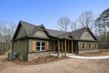 A modern house with dark green siding, stone accents, and wooden shutters stands in a wooded area. It has a gabled roof and inviting entrance.