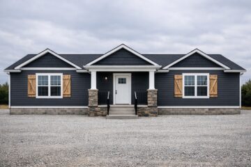 Single-story house with dark siding and white trim, featuring brick columns and wooden shutters. Cloudy sky and gravel foreground add a serene vibe.