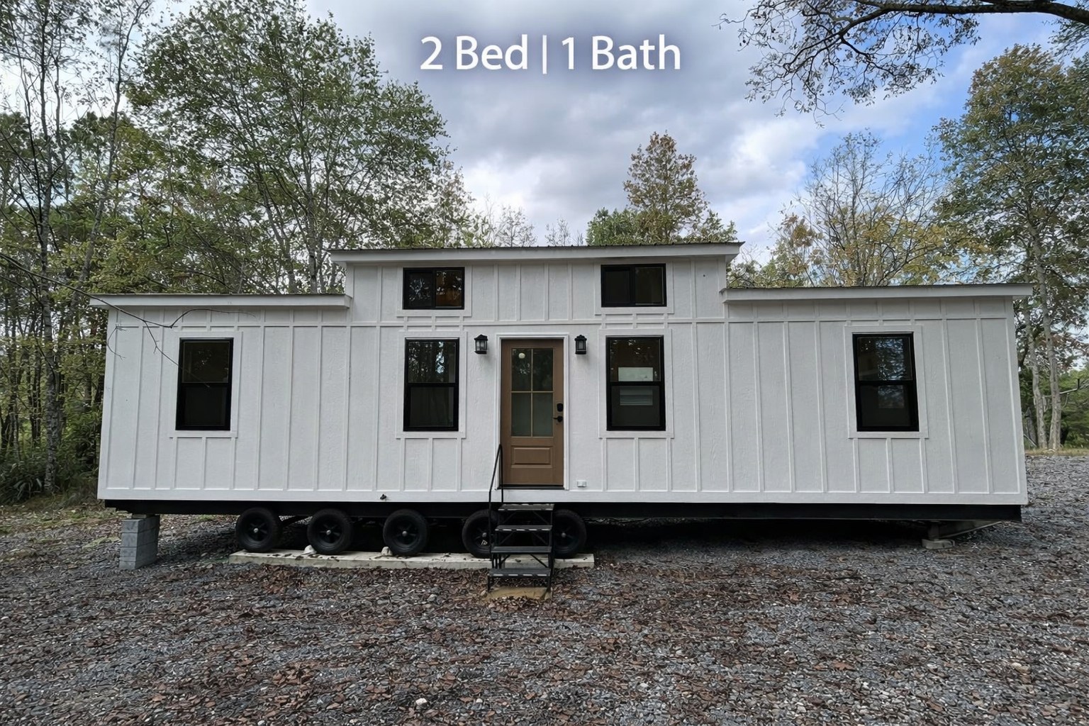 Small white mobile home titled "2 Bed | 1 Bath" on a gravel lot, surrounded by trees under a cloudy sky. Minimalist and compact design.