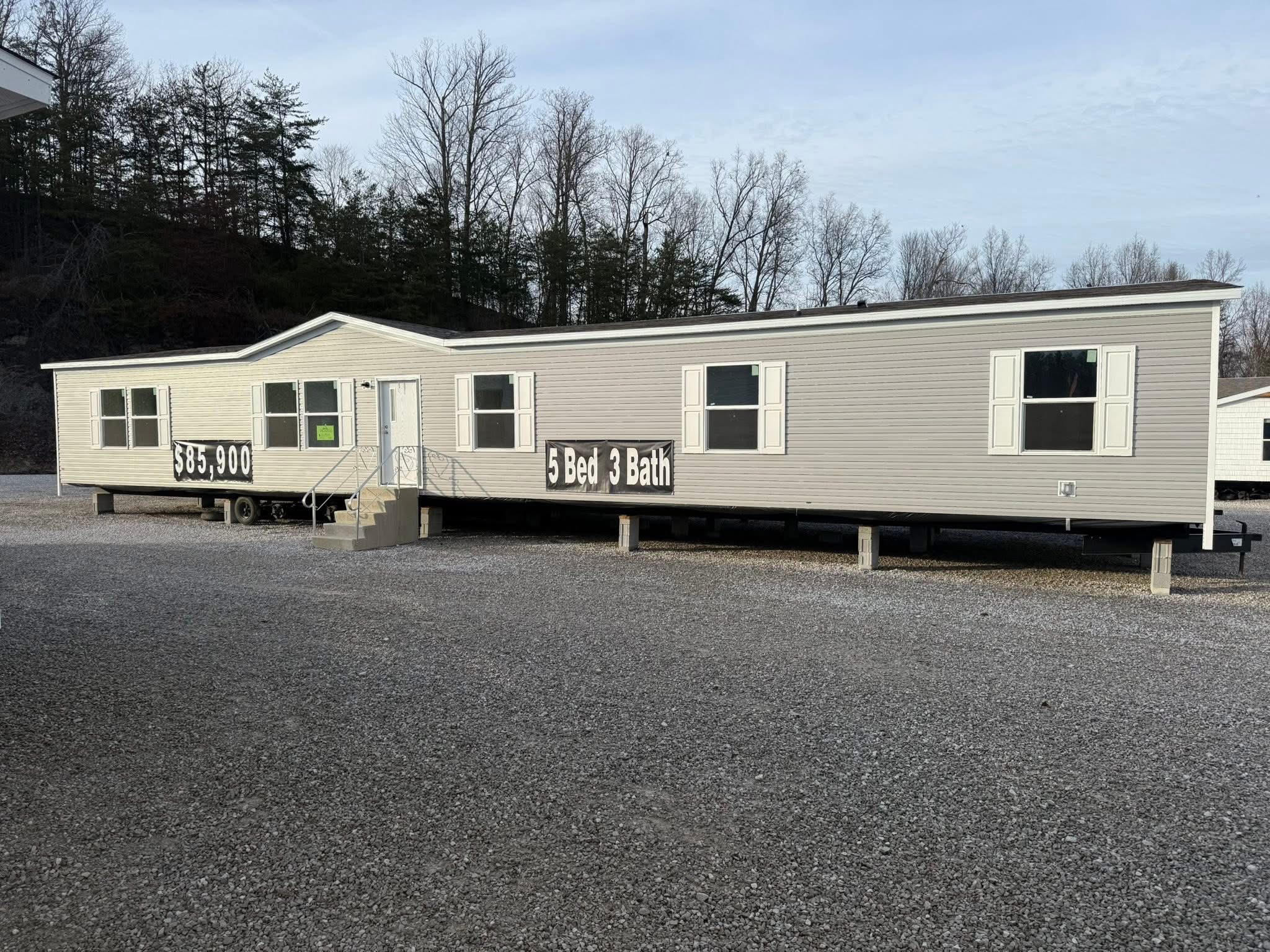 A beige mobile home on display in a gravel lot, featuring a "5 Bed 3 Bath" sign, priced at $35,900. Bare trees and overcast sky in the background.