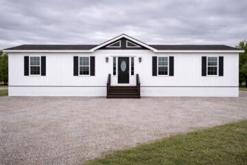 A white mobile home with black shutters and a centered dark door. It sits on a gravel driveway with cloudy skies overhead, evoking a calm rural setting.