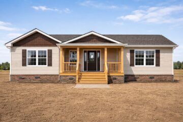 Single-story house with beige siding and brown accents, centered in an open, barren field under a clear blue sky. Features include a small porch and steps.
