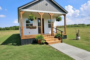 A small, charming gray cottage with a white door and wooden porch stands on a lush lawn. Hanging plants and a clear blue sky add a welcoming feel.