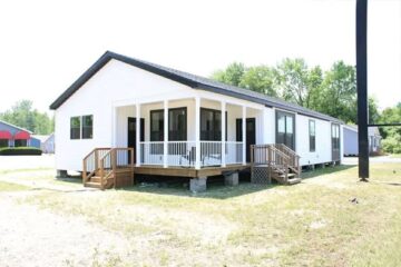 A white modular home with a black roof sits in a grassy area. It features a porch with railing and steps. The setting is bright and sunny, with trees in the background.
