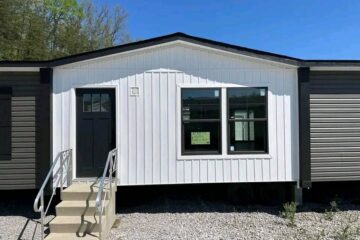 A modern one-story modular home with a white facade and gray sides. It has a glass-panel front door, three-step entry with metal railings, and a large window, set against a backdrop of clear blue sky and trees.